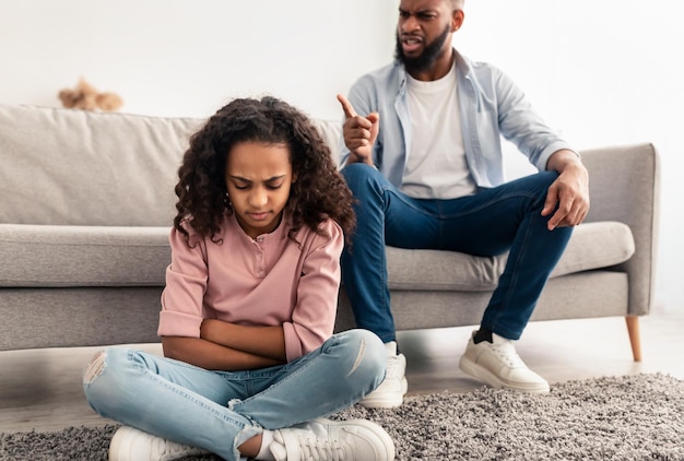 Naughty Kid. Portrait Of Angry Annoyed African American Father Scolding His Upset Little Daughter For Her Behaviour While She Sitting On The Floor At Home. Family Conflicts And Punishment Concept