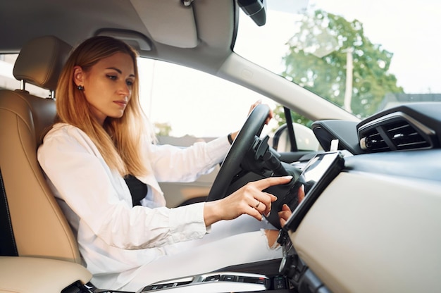 Navigating the vehicle's interface Young woman in white clothes is with her electric car at daytime