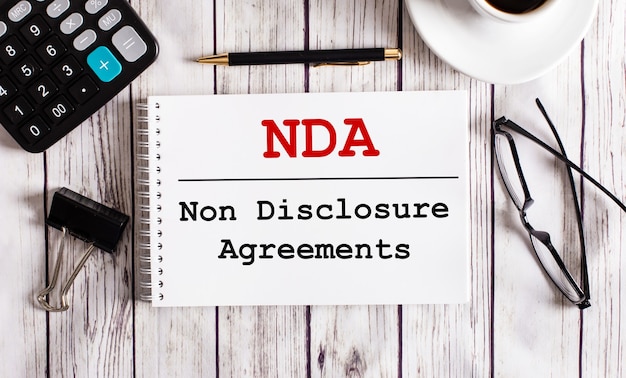 NDA Non Disclosure Agreements is written in a white notepad near a calculator, coffee, glasses and a pen. Business concept