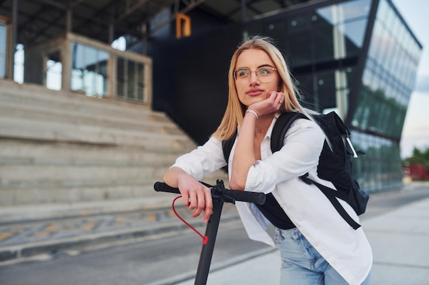 Near business building Beautiful blonde in casual clothes riding electric schooter outdoors at sunny daytime