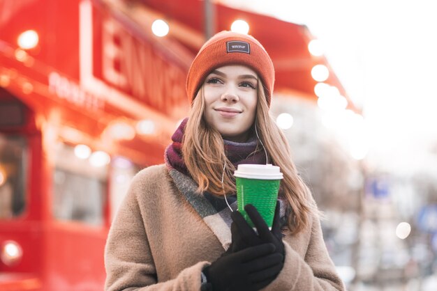 Near portrait teen girl in warm clothes standing outside on a cold spring day with a cup of coffee in his hands and looks aside in a great city bokeh