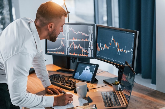 Near the table Young businessman in formal clothes is in office with multiple screens