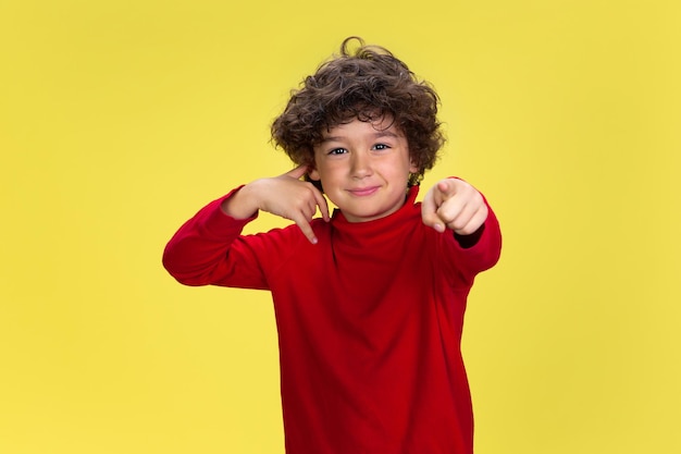 Need you to call Portrait of pretty young curly boy in red wear on yellow studio background Childhood expression education concept Preschooler with bright facial expression and sincere emotions