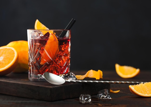 Negroni cocktail in crystal glass with orange slice and fresh raw oranges on chopping board with spoon on wooden background