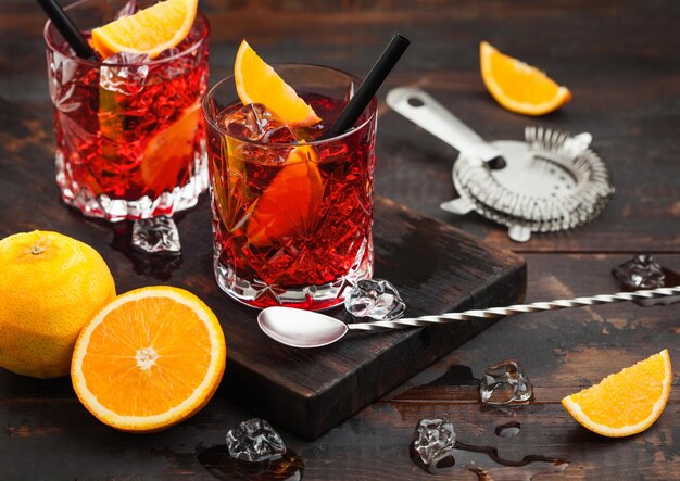 Negroni cocktail in crystal glasses with orange slice and fresh raw oranges with strainer and spoon on dark background.