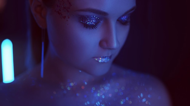 Photo neon girl portrait. glitter makeup art. woman with sparkling lips closed eyes in blue light.