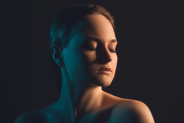 Neon light portrait. Beauty correction. Peaceful woman with bare shoulders closed eyes in blue glow isolated on dark copy space background. Aesthetic cosmetology. Skin rejuvenation.