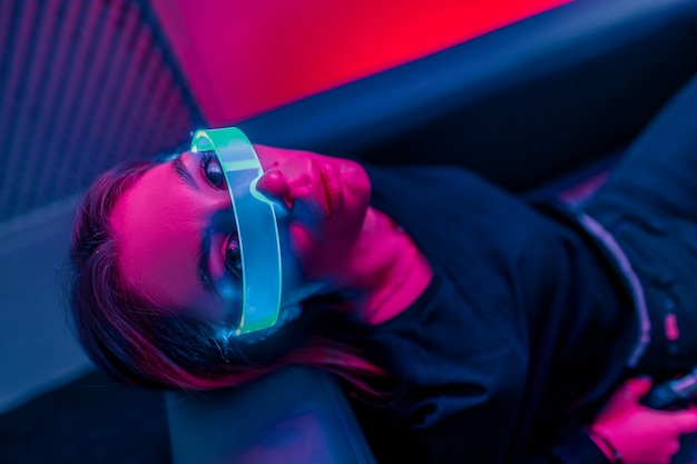 Neon purple light falls on a young woman in glowing glasses. Close-up. The photo has the effect of shush, grain. 