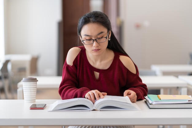 Nerdy asian girl reading textbook while preparing for exam or test in library reading room