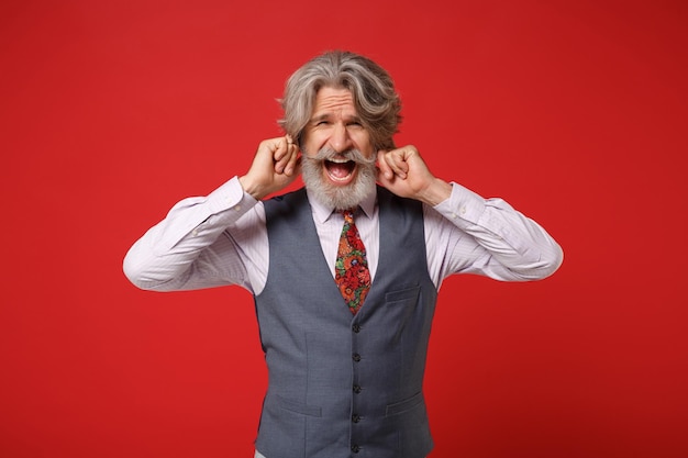 Nervous elderly gray-haired mustache bearded man in classic shirt vest and colorful tie isolated on red background. People lifestyle concept. Mock up copy space. Covering ears with fingers, screaming.