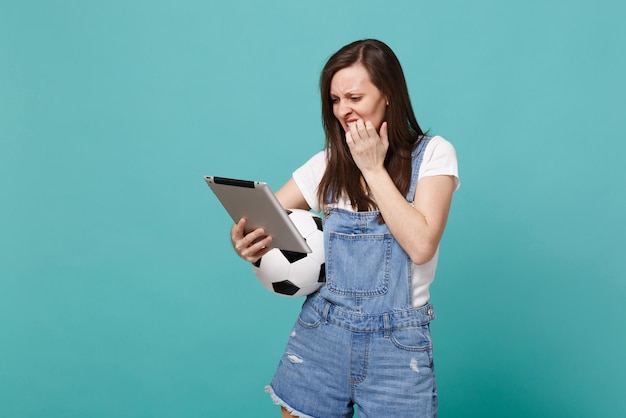 Nervous worried young woman football fan hold soccer ball, using tablet pc computer, gnawing nails isolated on blue turquoise wall background. People emotions, sport family leisure lifestyle concept.