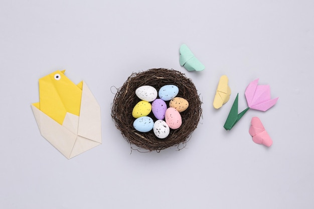 Nest with colored quail eggs and origami chicken tulip butterflies on gray background Easter composition