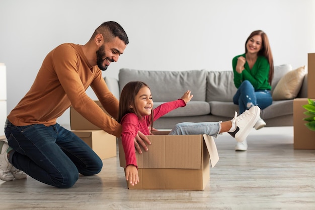 At New Apartment. Happy Middle Eastern family having fun and celebrating moving day, excited dad riding his small daughter in cardboard box, happy mom sitting on the couch in blurred background