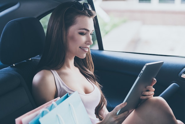 New collection in my favorite store! Beautiful young smiling woman looking at her digital tablet while sitting inside of the car with shopping bags laying near her