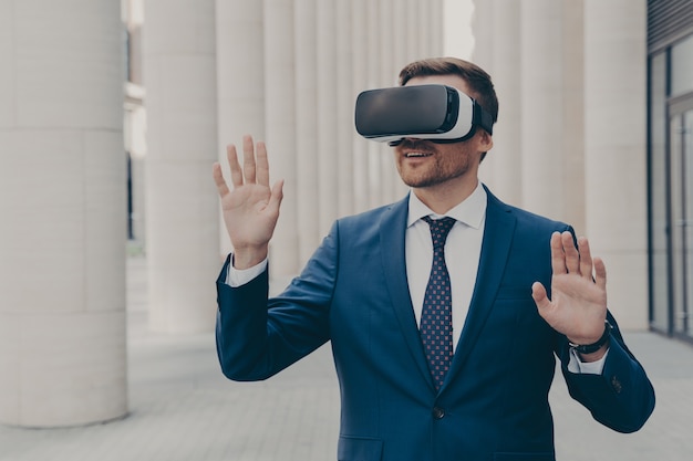 New cyberspace experience. Surprised male office worker dressed formally stands outdoors in 3d goggles and interacts with virtual interface, touching virtual objects, moving both hands in air