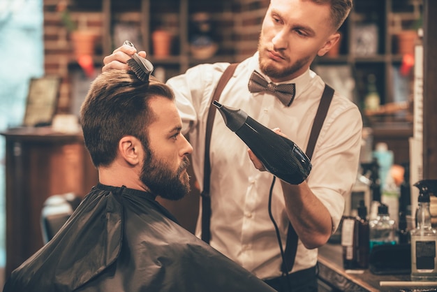 New hairstyle. Side view of young bearded man getting groomed at hairdresser with hair dryer