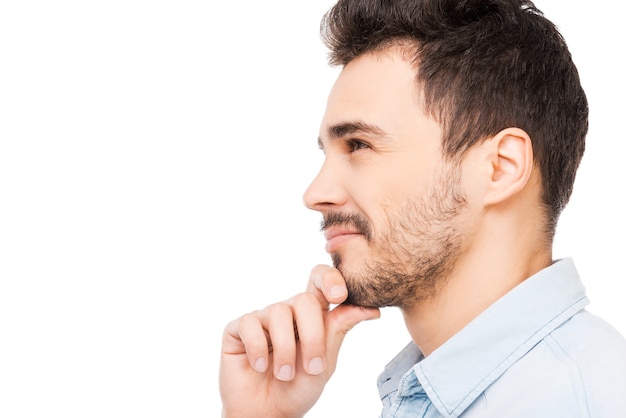 New ideas on my mind. Side view of thoughtful young man in shirt looking away and holding hand on chin while standing against white background