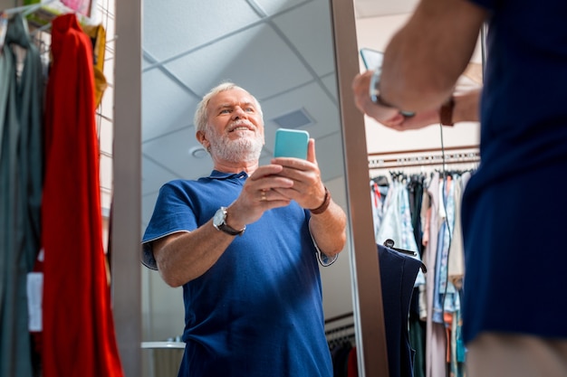 New image. Waist up of joyful grey haired man using mobile phone and making selfies in front of mirror while admiring his reflection