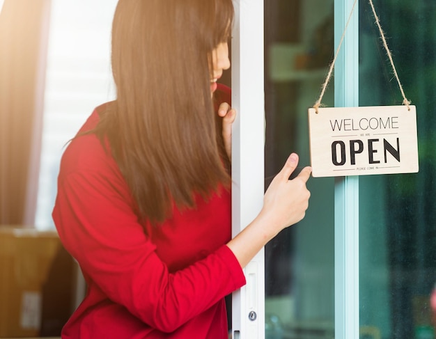 New normal, Asian young woman glad and smiling point finger to notice sign wood board label "WELCOME OPEN" hang through glass door front shop, Business turning open after coronavirus pandemic disease