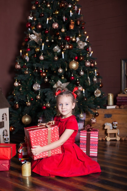 New Year 2020! Merry Christmas, happy holidays! Little girl holds a gift, smiles, rejoices and awaits Christmas, New Year and holiday. A cozy house, a burning fireplace and a Christmas tree. Winter