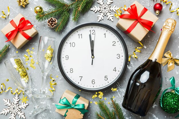 New year celebration concept with a bottle of champagne and two glasses toasting Christmas gift box alarm clock and fir tree branch on colored table Top view Copy space