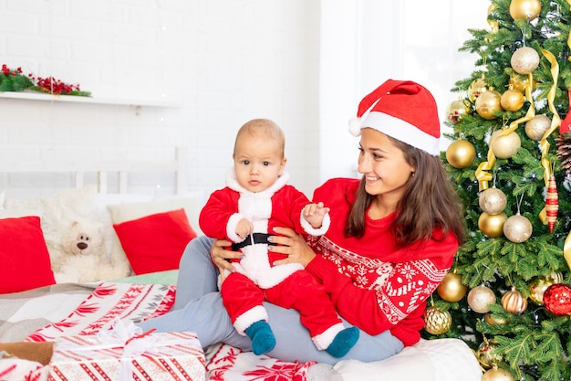 New Year or Christmas, a young mother with a baby on the bed at home by the Christmas tree in a Santa Claus costume hugging and waiting for the holiday smiling