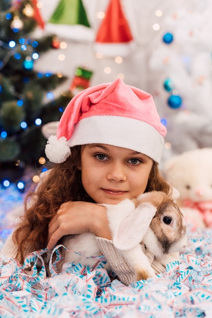 New Year a girl in a pink santa hat lies near a christmas tree and strokes a rabbit