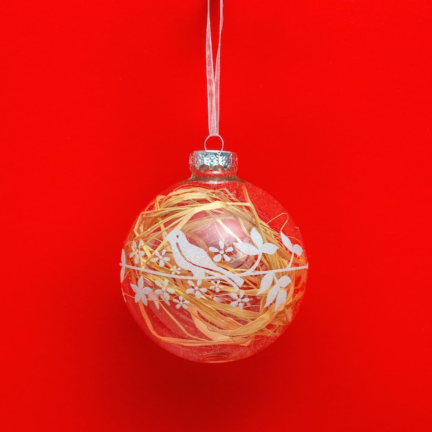 New Year glass ball transparent with painted bird and natural material inside on red paper background. Creative holiday christmas concept.