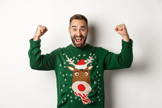 New Year, holidays and celebration. Relieved and excited man in Christmas sweater, raising hands up and celebrating, triumphing, standing over white background.