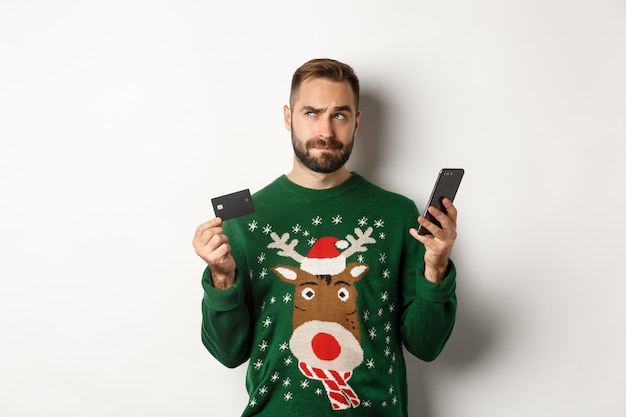 New year online shopping and christmas concept thoughtful man using mobile phone and credit card thi...