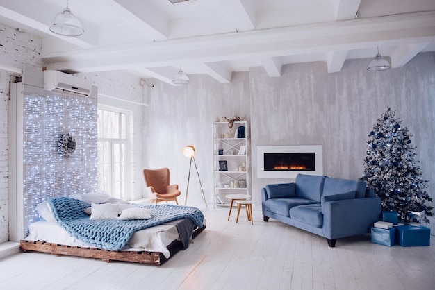 New Year's interior of the apartment. Big Windows, blue sofa, artificial fireplace, bed, lights, white and blue Christmas tree, armchair and stylish floor lamp. Loft style. Concrete wall. Cozy house.