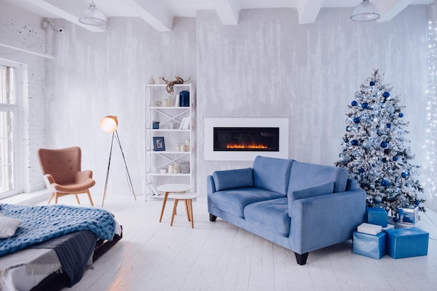 New Year's interior of the apartment. Big Windows, blue sofa, artificial fireplace, bed, lights, white and blue Christmas tree, armchair and stylish floor lamp. Loft style. Concrete wall. Cozy house.