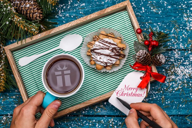 New Year's photo of tea with picture of gift, cake on table with spruce branch, person writing wishes on postcard