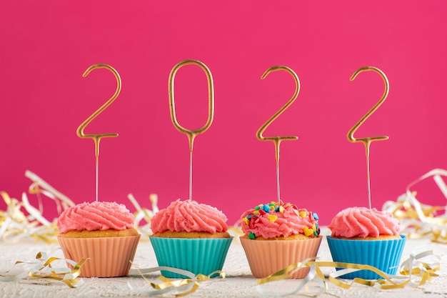 New Years background. Holiday cupcakes with numbers 2022 surrounded by New Years tinsel on pink background. Copy space.