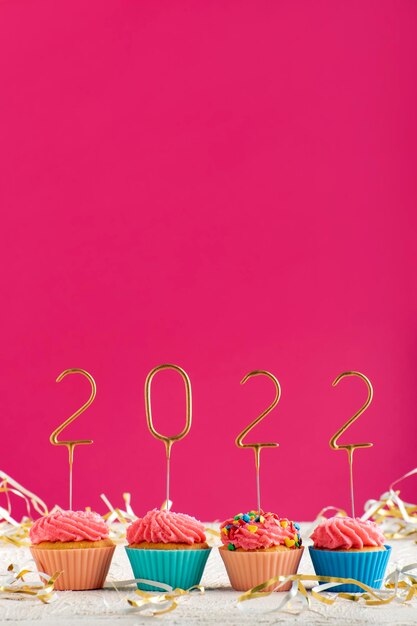 New Years background. Holiday cupcakes with numbers 2022 surrounded by New Years tinsel on pink background. Vertical frame. Copy space.