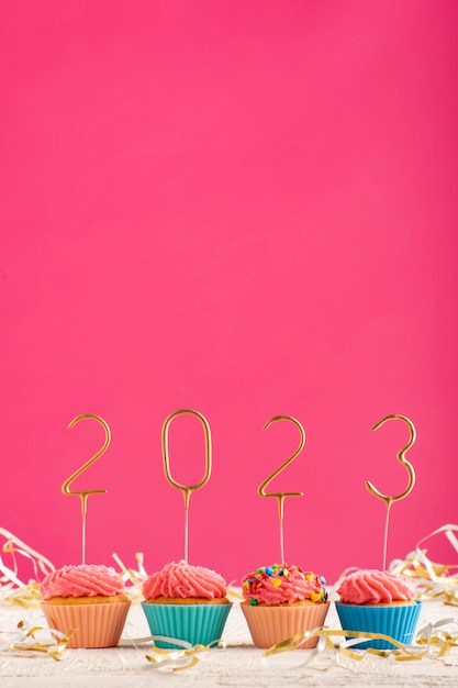 New Years background Holiday cupcakes with numbers 2023 surrounded by New Years tinsel on pink background Vertical frame Copy space