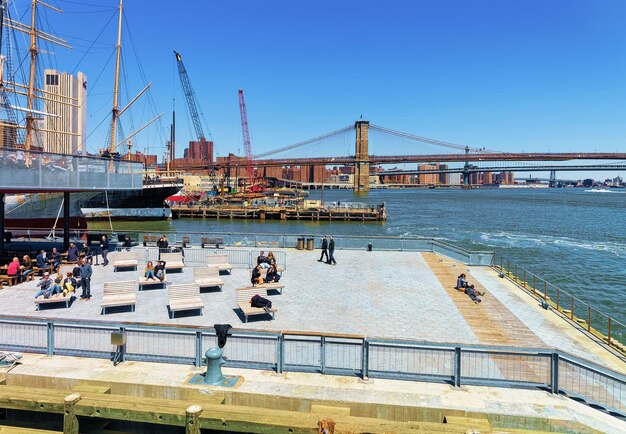 New York, USA - April 25, 2015: Tourists relax on sunbeds at Pier in Lower Manhattan, New York. View from Ferry on Pier, Brooklyn Bridge and Manhattan Bridge and Brooklyn, the USA, East River