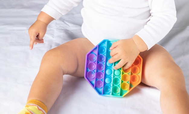 Newborn baby sits on the bed playing trendy flexible silicone multicolored toy pop it Selective focus on anti stress simple dimple toy in children's hands