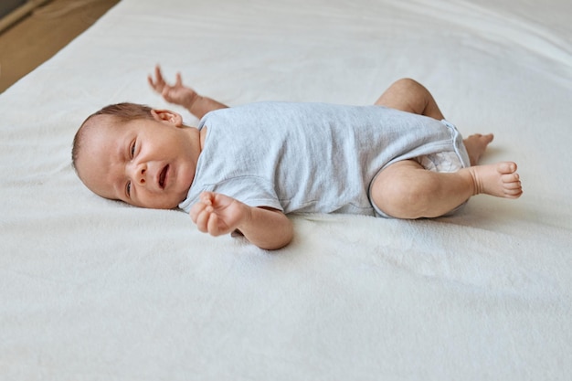 Photo newborn baby wearing bodysuit crying while laying on white sheet having childish tantrums suffering colic and abdominal pain infants needs medicine help or mother attention