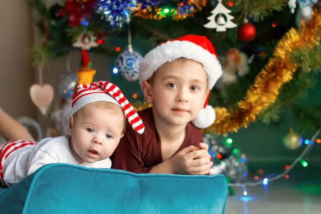 Newborn baby with older brother posing against the background of Christmas tree. Portrait of two lovely brothers in santa hats. Christmas time. Family concept.