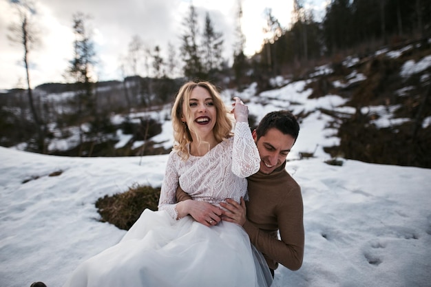 Newlywed groom and bride playing in snow husband hugging his wife in winter forest park family outdoor activities