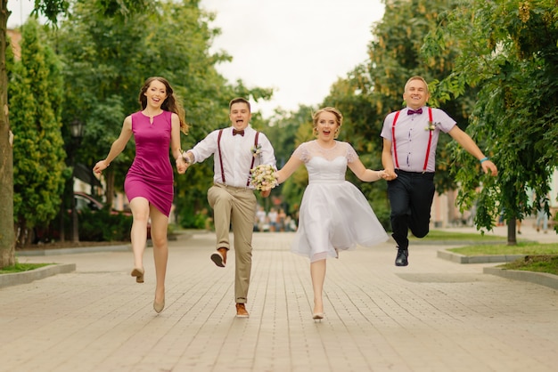 Newlyweds are have fun at wedding day together with their friends, holding hands they run along the street.