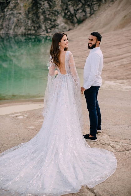 Newlyweds are standing near a beautiful lake. Middle Eastern groom and Caucasian bride hold hands on the beach. Back view.