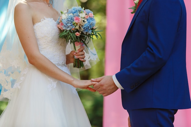 Newlyweds holding hands at sunny wedding day.  Bride with a beautiful bouquet of pink and white  flowers and groom in blue suit .wedding ceremony outdoors.wedding. romantic moment close up