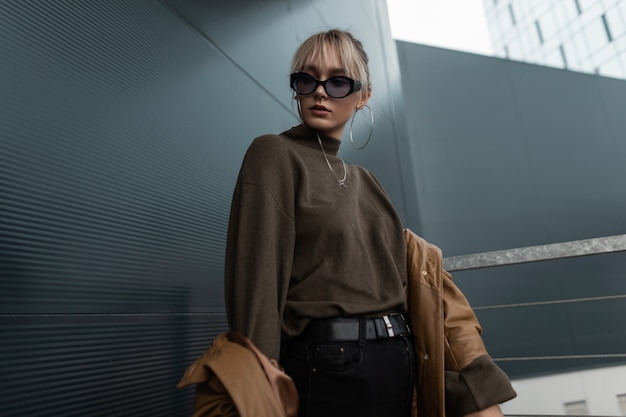 Nice american model girl in youth clothes with a leather brown jacket, black jeans and sweater in vinrage sunglasses posing in the city