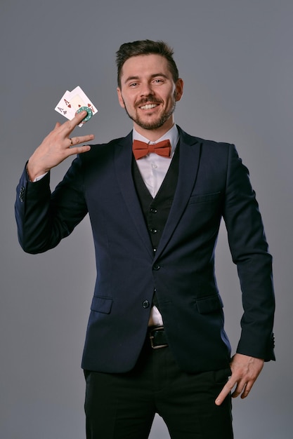 Nice brunet guy in black classic suit and red bow-tie is smiling, showing two playing cards and chips while posing against gray studio background. Gambling entertainment, poker, casino. Close-up.