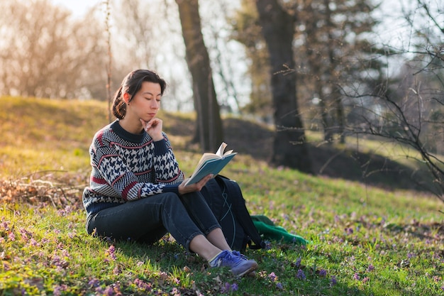 Nice brunette girl is sitting on the grass in the park and reading a book.