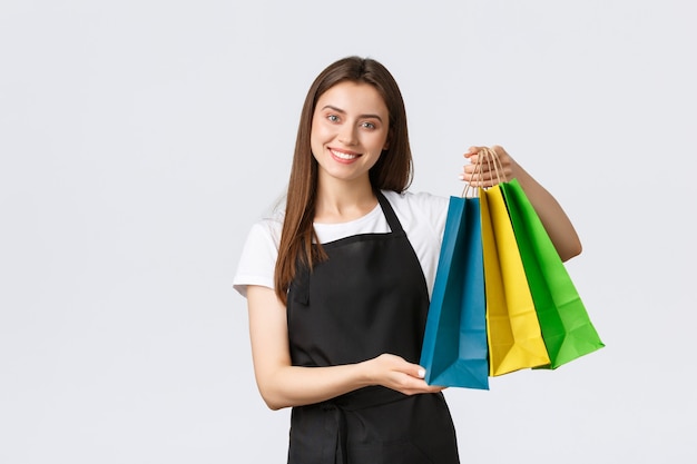 Nice charming smiling female cashier in black apron give customer their bags with purchased goods and wishing nice day. Saleswoman working over counter, standing white background.