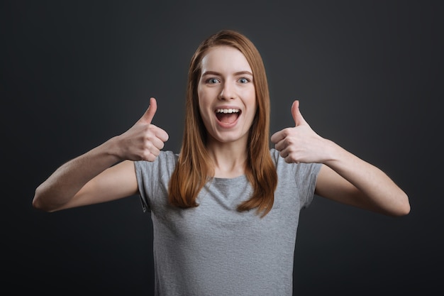 Nice choice. Encouraging expressive gorgeous girl showing the sign of approval while posing for a photographer and standing isolated on grey background