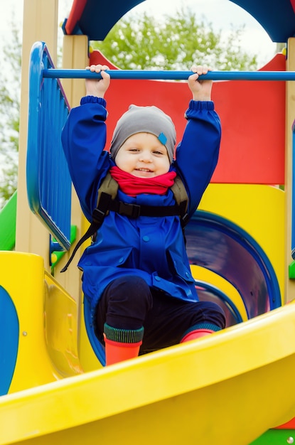 Nice cute boy of two years in a blue jacket and red scarf is playing on a bright playground